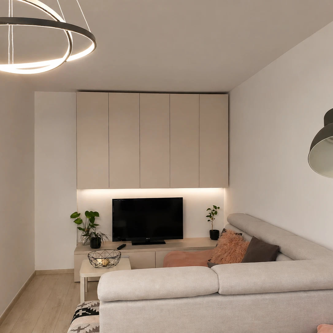 Wohnraum einer kleinen Wohnung: ein helles Ecksofa mit rosé Kissen, eine cremefarbene raumhohe Schrankzeile über einem niedrigen, von unten beleuchteten Medienmöbel, darüber eine Ringleuchte und rechts eine dunkle, geneigte Stehlampe.