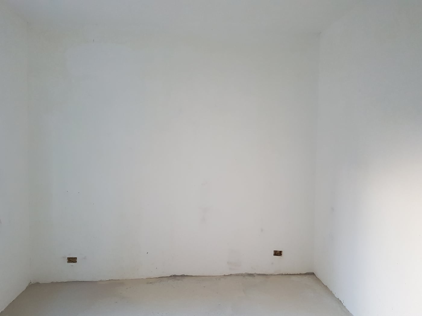 The same room stripped back: bare white walls, a raw screed floor and two sockets chased into the plaster.